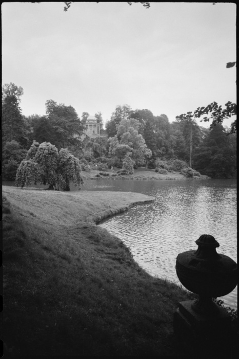 Temple of Apollo, Stourhead, Wiltshire, c1955 1980. Creator: Ursula Clark. Temple of Apollo, Stourhead, Wiltshire, c1955 1980. View of the garden lake in the grounds of Stourhead House, with the Temple of Apollo in the distance and a stone urn in the foreground, seen from a bank on the east of the lake and north of the temple. The image shows a bank of the lake in the foreground with a steep bend in the bank, creating a wide body of water in front of the hill that the temple sits on. At the base of the hill are a few people on a path and foliage that extends to the top of the hill. The Temple of Apollo is in a clearing at the apex and is a circular building on a plinth with eight steps, built in 1765. The design is based on a temple in Wood s Ruins of Baalbec, which was published in 1757. The temple is surrounded by Corinthian columns that support scalloped mouldings and a domed roof. Each bay sround the temple has a niche with circular oculus which had lead statues that were moved to the house in 1903 1904. The house and grounds are now cared for by the National Trust.