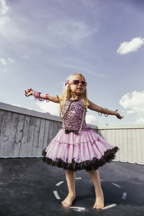 A young girl all dressed up with blond curly hair, sunglasses and jewelry standing on a trampoline in the backyard and showing off; Spruce Grove, Alberta, Canada