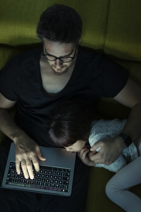 Father using laptop on couch at night with daughter  lying next to him Father using laptop on couch at night with daughter  lying next to him