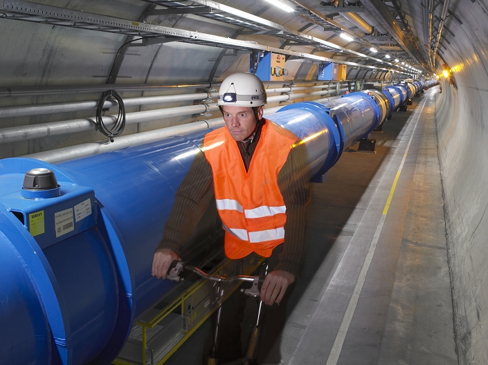 LHC tunnel, composite image. Safety supervisor riding a bicycle along the LHC (large hadron collider) tunnel at CERN (the European particle physics laboratory) near Geneva, Switzerland. The LHC is a 27-kilometre-long underground ring of superconducting magnets housed in this pipe-like structure (blue), or cryostat. The cryostat is cooled by liquid helium to keep it at an operating temperature just above absolute zero. It will accelerate two counter-rotating beam of protons to an energy of 7 tera electron volts (TeV) and then bring them to collide head on. Several detectors are being built around the LHC to detect the various particles produced by the collision. A pilot run of the LHC is scheduled for summer 2007.