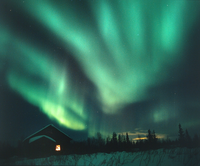Aurora borealis over a cabin. This coloured light display, also called the northern lights, is observed in the night sky at high latitudes. It occurs when charged and highly-energetic particles from the Sun (the solar wind) are drawn by Earth's magnetic field to the northern and southern polar regions. Hundreds of kilometres up, they collide with the upper atmosphere, causing the excitation of atoms and molecules which leads to the emission of light. Oxygen atoms (rather than molecules) are the most abundant components of the atmosphere at these heights. It is their excitation that results in green light. Photographed in March 2001, near Yellowknife, Northwestern Territories, Canada.