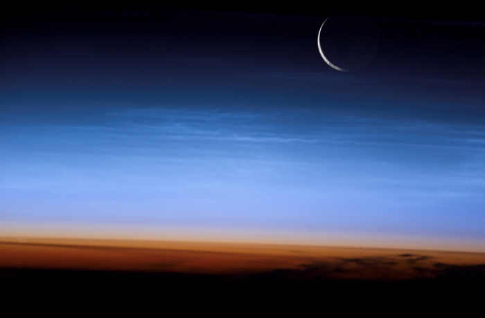 Noctilucent clouds, seen from the International Space Station (ISS). These clouds (white streaks across centre) are very thin and form high above the Earth at heights of 75-90 kilometres. Because they are so thin and high, they can only be seen at twilight, shining in the glow of the setting Sun, and only at high and low latitudes. The lower three layers of the atmosphere (from bottom) are the troposphere (orange, 15 kilometres thick), and the stratosphere and mesosphere (both pale blue & 35 kilometres thick). Above these, the atmosphere fades into the darkness of space. A crescent Moon is also seen. The ISS orbits some 380 kilometres high. Photographed on 27 July 2003, over Mongolia.