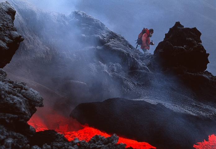 . Volcanologist wearing a gas mask and holding a camcorder as he stands next to a lava flow on the slopes of the volcano Mount Etna. Lava is molten rock that is produced by volcanoes as an upwelling of the magma found beneath the Earth's crust. The lava can range in temperature from 475 degrees Celsius (dim red) to 1150 degrees Celsius (white). It solidifies to form the black volcanic rock surrounding the flow. Other dangers on the slopes of a volcano include poisonous gases such as hydrogen sulphide, and lava bombs, chunks of molten rock ejected by the volcano. Mount Etna, standing 3323 metres tall on the Italian island of Sicily, is the largest active volcano in Europe. MODEL RELEASED
