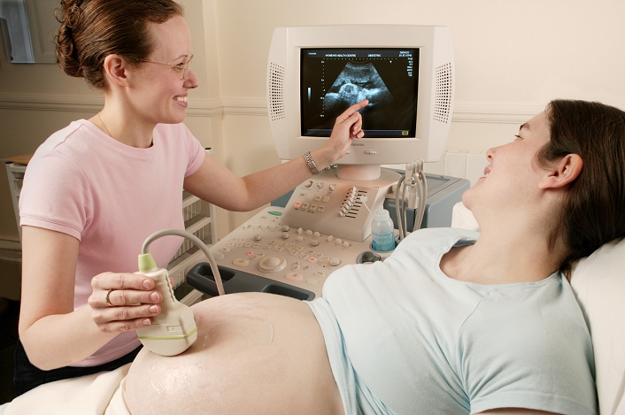 . Pregnancy ultrasound. Sonographer pointing out parts of a expectant mother's foetus to her on an ultrasound scan. The sonographer is holding a transducer to scan the woman's abdomen. The transducer emits high- frequency sound waves and detects the reflected echoes. The results are used to build an image of the developing foetus in the womb, which is displayed on a monitor. This is a safe, non- invasive procedure used routinely to assess the growth and health of the developing foetus. MODEL RELEASED