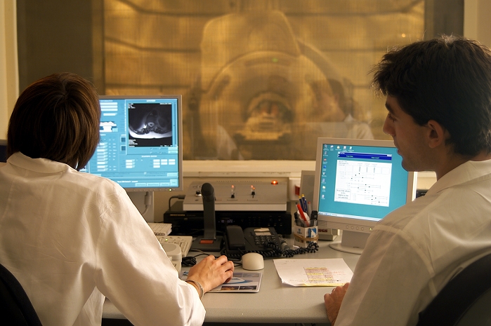 . MRI scanning. Radiologists looking at a magnetic resonance imaging (MRI) scan (screen at left) of the right knee of a 60-year-old woman. The patient and the MRI scanner are seen through the window in the background. The patient, only partially seen, enters the scanner through the central hole. MRI scanning uses strong magnetic fields to produce radio emissions from hydrogen atoms in the body. MRI scanning is used to produce cross-sectional body images, and is most useful for imaging soft tissues. Photographed at the American Hospital of Paris, France. MODEL RELEASED