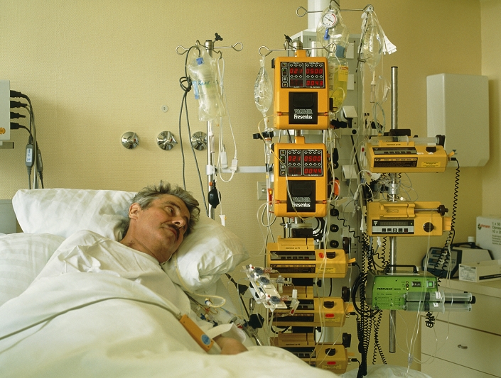 POSED BY MODEL. Cardiac intensive care. Male patient recovering in a cardiac intensive care unit (ICU) after a heart transplant. The devices at right control the delivery of fluids and drugs from intravenous drip bags. Intensive care may also be required following a serious infection, an accident or a heart attack.