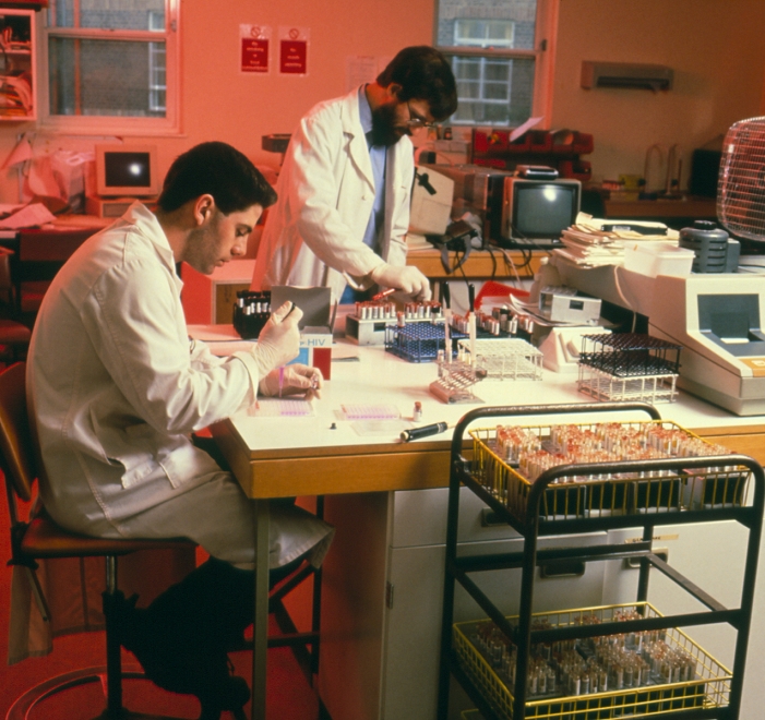 . Scientists screening blood at the North London Blood Transfusion Centre. The man on left is performing a Fuji-Rebio test for antibodies to HIV-1 (Human Immunodeficiency Virus type 1), a causative agent of AIDS. Human serum is diluted then placed in both a test and a control well. Gelatine particles & HIV antigens are added to the test well but only gelatine particles to the control well. Any HIV-antibodies present in serum agglutinate with antigens in the test well & provoke the appearance of a lattice in the bottom of the well. A small 'button' of particles would be the appearance of a normal control sample. MODEL RELEASED