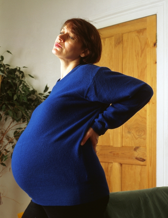 . Backache in pregnancy. Heavily pregnant woman stands holding her back in an expression of pain. Lower back pain is common towards the end of pregnancy. MODEL RELEASED