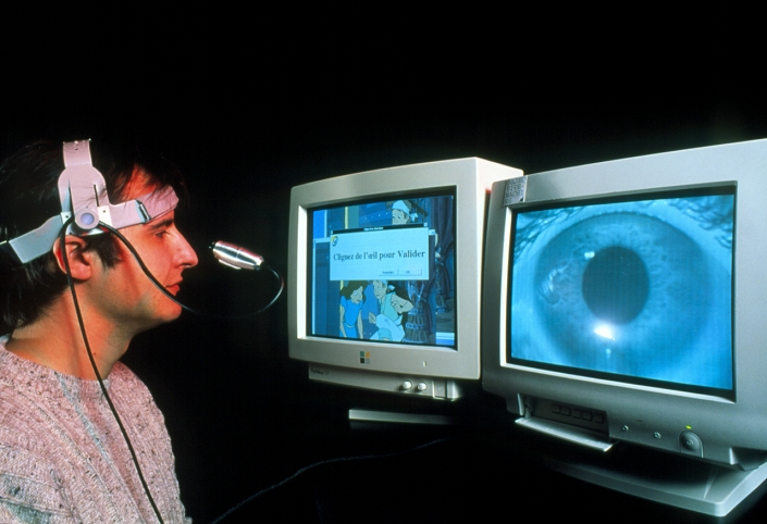 Interactive computer. Researcher playing a game on an eye access interactive computer system. He has a small camera mounted on his head which tracks the movements of his eye and translates it into the movements of a pointer on the screen. The camera's view is seen on the monitor at lower right. The text on the screen at centre informs the man (in French) to blink to validate his command. This system could be used to help severely disabled people who cannot use a mouse or keyboard to interact with computers. As well as games, facilities such as word processors, voice synthesisers and even software to control devices in the home could be operated using this system.