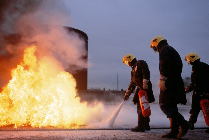 Firefighters putting out a fire during a training exercise. The firefighters are wearing protective clothing, which includes fire-proof jackets and trousers, and helmets. The firefighters are using a fire extinguisher to spray foam at the base of a fire. This is a simulation of a fire at a petroleum works. Photographed in France.