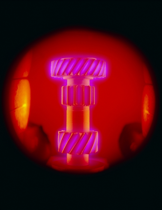 A transmission gear undergoing a surface hardening process called plasma carburisation within a plasma chamber. Ions from the plasma dissolve into the steel (which is given a negative charge to attract them) thus forming a new, harder, alloy at the surface. The plasma contains the correct mixture of elements necessary to produce the desired characteristics in the new alloy. This photo, taken through the inspection window of the sealed chamber, shows the part fluorescing. The energy responsible for this is evolved when the excited plasma ions recombine with electrons in the steel thus achieving a lower energy state.