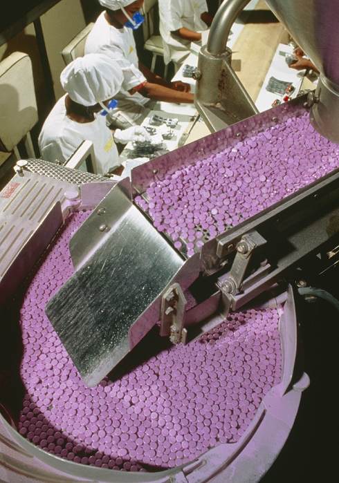 Drug packaging. View of a feed hopper of a tablet packaging machine. The tablets are packaged in plastic and foil sheets by the machine. The workers in the background check the foil packaging, then place the sheets into boxes. Photographed in Nigeria.