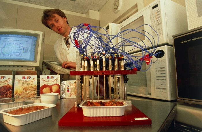 Food additives research. Researcher analysing food after it has been cooked in a microwave oven as part of research into the taste and aroma of different foods. The probes measure the temperature at different points within the food and have produced the contours on the heat map on the computer screen at upper left. Heating the food mobilises the chemicals which cause the taste and aroma of food. The taste and smell will be noted by researchers before the natural chemicals which cause them are separated out and identified. These chemicals can be used to make artificial flavours and aromas for addition to food. Photographed in France.