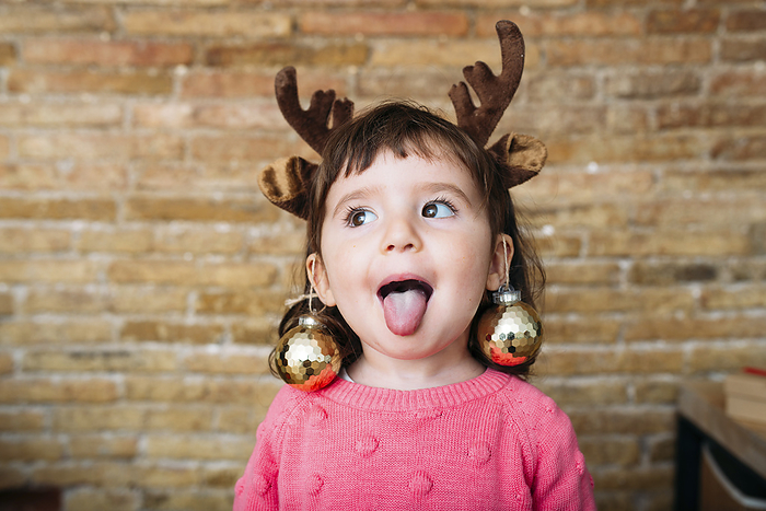 AbrilRoman.jpg Portrait of toddler girl sticking out tongue wearing reindeer antlers headband and Christmas baubles