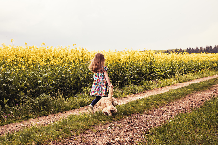Girl walking alone with teddy and backpack on a field way