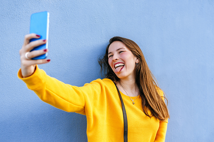 Woman holding a cell phone Happy young woman taking a selfie at a wall sticking out tongue