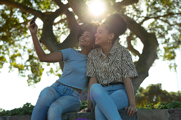 Front view close up of two young smiling adult mixed race sisters sitting on a wall in an urban park, using a smartphone and taking selfies, backlit with lens flare