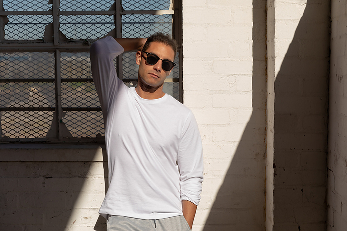 Portrait of a young Caucasian man wearing sunglasses standing against a wall in a shaft of sunlight with one hand behind his head, looking to camera at an abandoned warehouse