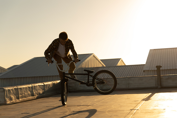 Side view of a young Caucasian man riding a BMX bike and doing tricks on the rooftop of an abandoned warehouse, backlit by the setting sun Side view of a young Caucasian man riding a BMX bike and doing tricks on the rooftop of an abandoned warehouse, backlit by the setting sun