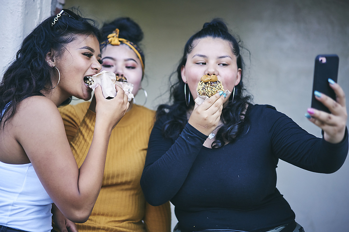 Young woman and teenage sisters posing for selfie while eating cakes Young woman and teenage sisters posing for selfie while eating cakes