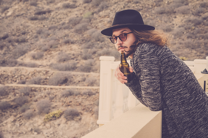 long hair beautiful young man drinking beer outdoor on the rooftop terrace with mountain and nature in background. vingtage retro filter. black hat and eyeglasses, Photo by Fabio and Simona