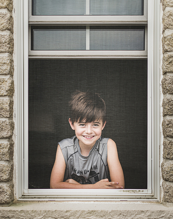 Young boy looking out through the screen of an open window.