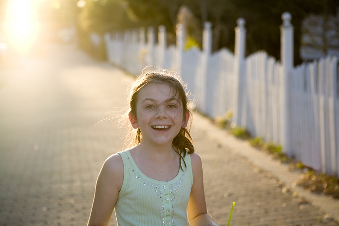 A freckled girl is smiling with a picket fence and sun in the background.