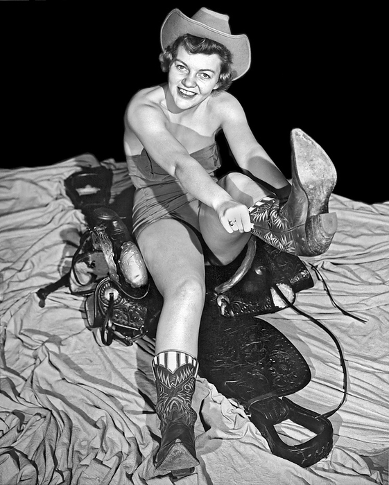Texas:  1954. 
A pretty young woman sitting on a saddle while wearing a bathing suit and a Stetson hat pulls on her cowboy boots.