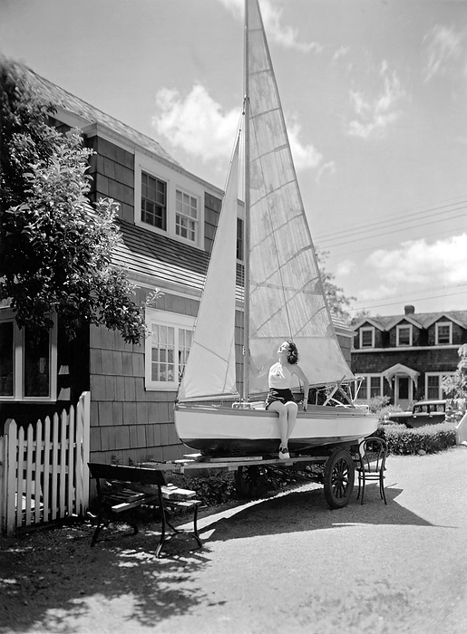 United States:   c. 1933.
A young woman catches some rays from the sun on the deck of a sailboat that is on a trailer in the driveway