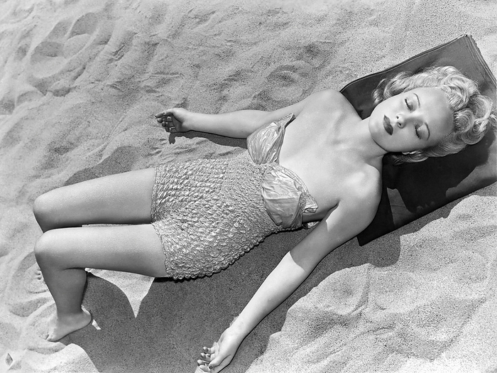 United States:  c. 1958.
A pretty woman on the beach in a one piece bathing suit closes her eyes  and soaks in the sun's rays.