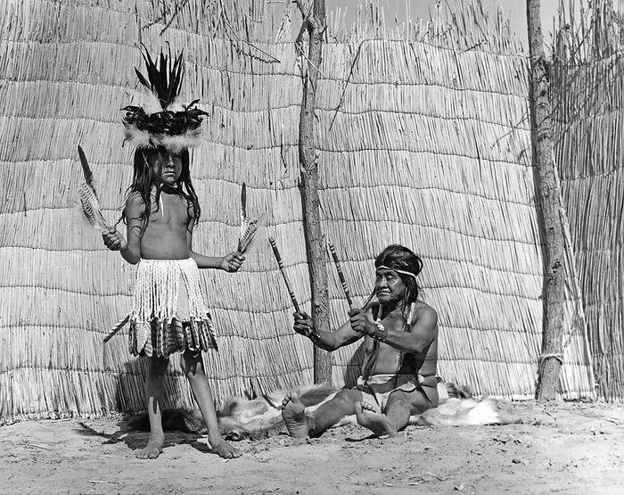 California:  c. 1930
Medicine men taught their songs and dances to young boys chosen to carry on the traditions of the tribe. This Yokut medicine man is playing his split clap sticks by shaking them in time to his song. The dancing costume is a skirt of eagle down tipped with red-tailed hawk feathers. The hat is a basket decorated with eagle down, and raven and magpie feathers.