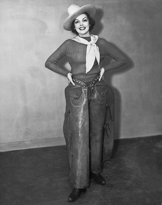 United States:  c. 1940
An attractive woman wearing a cowboy hat, kerchief, boots, and chaps stands smiling with her hands on her hips.
