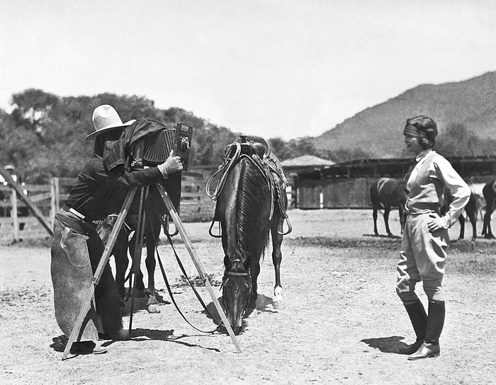 United States:  c.  1924
A cowboy with a camera on a tripod takes a photograph of a pretty young woman.