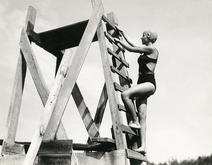 United States:   c. 1931
A young woman climbing up to the top of the wooden high dive platform.