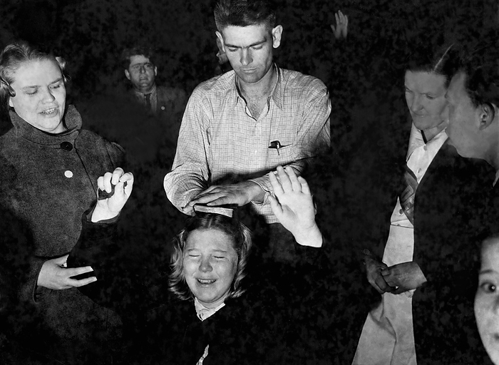 United States:  c. 1948
A man places a bible on the head of an ecstatic kneeling worshipper responding to the call of the preacher at a religious tent revival meeting.