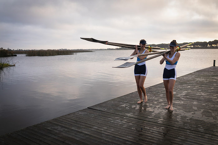 Front view of two Caucasian women from a rowing team carrying oars on their shoudlers and walking along a jetty on the river at sunrise, talking and smiling