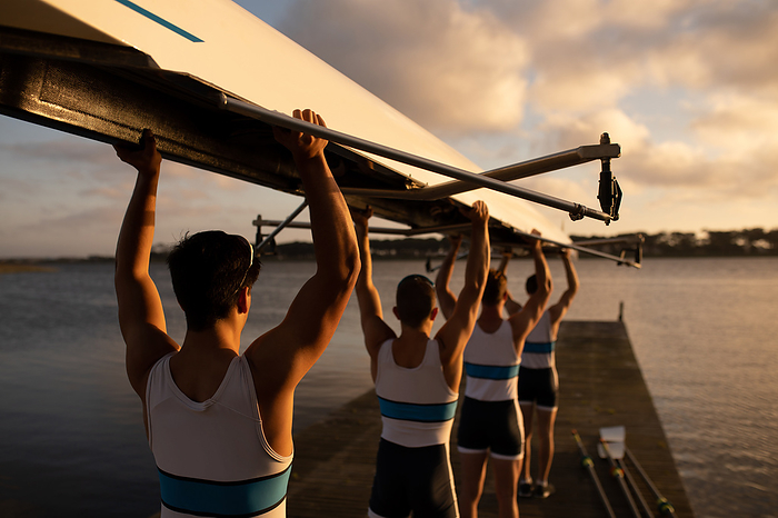 Rear view of a rowing team of four Caucasian men carrying a boat above their heads with arms raised, walking along a jetty on the river at sunset