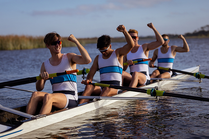Side view of a rowing team of four Caucasian men training and rowing on the river, sitting in a rowing boat, cheering with their arms raised, enjoying the training