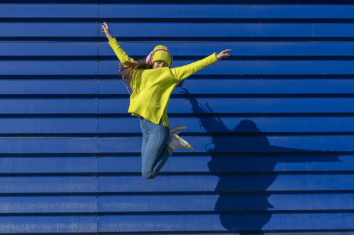 Teenage girl jumping in the air in front of blue background listening music with headphones