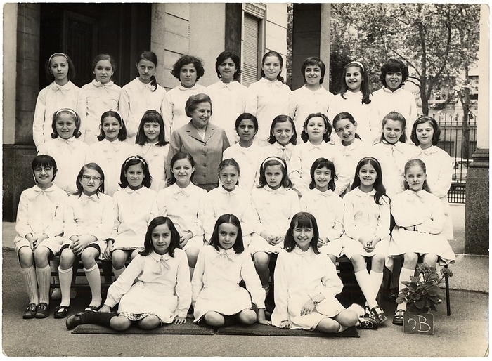 FEMININE PHILOGY SCHOOL SCHOOL Photography souvenir of a female elementary class: 5B. The students wear all the classic white apron distinguished by a light ribbon. In the first row in the middle two twins. The girls smile all or almost, just like the teacher who smiles but who, unlike her pupils, does not look in the car. The weather is cheerful, even a little girl babe takes out her tongue in a mocking expression. The single teacher alone managed the entire class of 30 students, a case not uncommon in the baby boom years, with the schooling set for the achievement of the eighth grade diploma or  alternatively  at the age of 14 years. The numerous successive reforms of the school have begun a period of experimentation that has led to an evolution of teaching, also decreasing the number of students per class and adding one or more colleagues alongside the owner, in order to guarantee to all schoolchildren opportunities to study, and to discourage school leaving in accordance with the increase in the age of the obligation. Group photo in the schoolyard, Milan 1971.