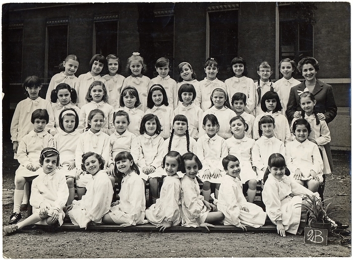 FEMININE PHILOGY SCHOOL SCHOOL Photography souvenir of a female elementary class: 2B. The students wear all the classic white apron distinguished by a light ribbon. In the center of the first row a pair of twins appropriately posed by the photographer in order to create a choreographic mirror effect. The single teacher, posing as smiling as all the girls, managed the entire class of more than 35 students, an event not rare in the baby boom years, with the schooling set for the achievement of the diploma of eighth grade or  alternatively   at the age of 14. The numerous successive reforms of the school have begun a period of experimentation that has led to an evolution of teaching, also decreasing the number of students per class and adding one or more colleagues alongside the owner, in order to guarantee to all schoolchildren opportunities to study, and to discourage school leaving in accordance with the increase in the age of the obligation. Group photo in the schoolyard, Milan 1968.