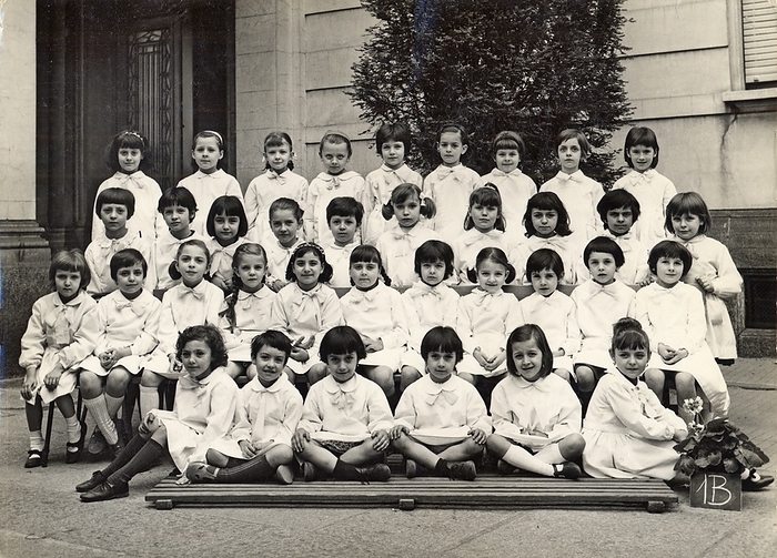 FEMININE PHILOGY SCHOOL SCHOOL Photography souvenir of a female elementary class: 1B. The students wear all the classic white apron distinguished by a light ribbon. In the center of the first row a couple of twins. The single teacher, not present in the photo, managed alone the entire class made up of more than 35 students, a case not rare in the baby boom years, with the schooling fixed to the achievement of the diploma of third grade or  alternatively  to completion of 14 years of age. The numerous successive reforms of the school have begun a period of experimentation that has led to an evolution of teaching, also decreasing the number of students per class and adding one or more colleagues alongside the owner, in order to guarantee to all schoolchildren opportunities to study, and to discourage school leaving in accordance with the increase in the age of the obligation. Group photo in the schoolyard, Milan 1967.