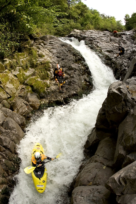Overhead view of a kayaker going down a narrow and steep rapid with other kayakers watching.