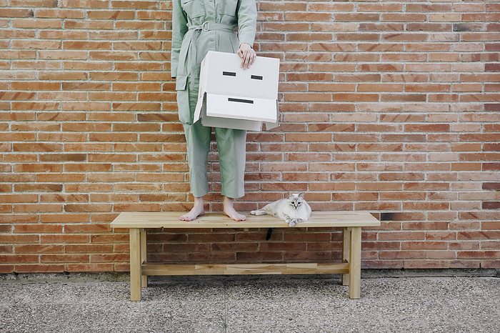 Woman standing on bench with cat in front of brick wall holding  cardboard box with sad face