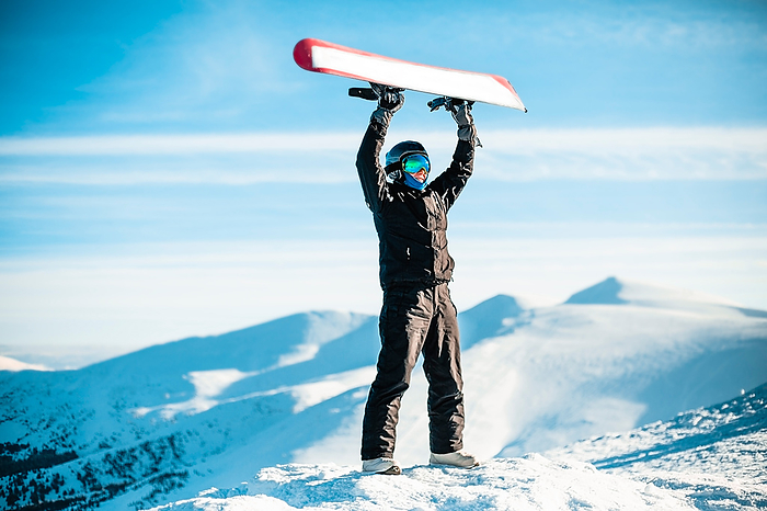 A person wearing a black ski suit, helmet and goggles on top of a mountain holding a red snowboard above their head. A person wearing a black ski suit, helmet and goggles on top of a mountain holding a red snowboard above their head.