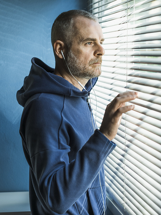 Pensive man with earbuds looking out of venetian blind window