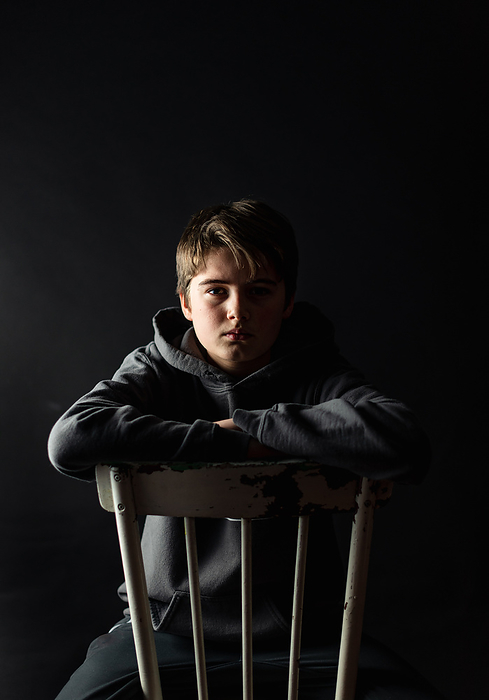 Low key portrait of adolescent boy sitting on a chair in a dark room., Kingston, ON, Canada