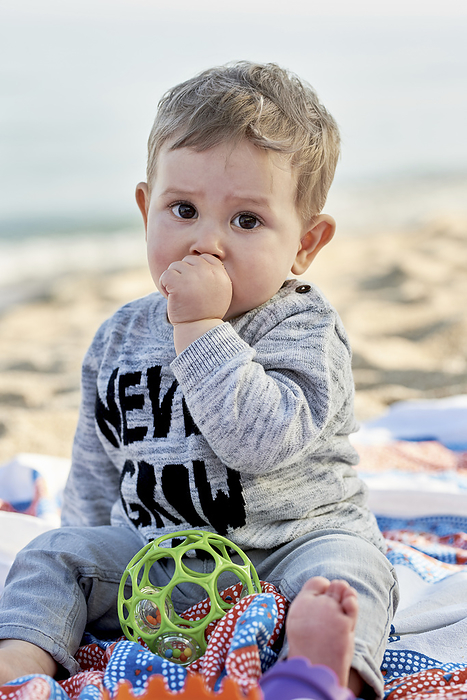 Cute boy sucking thumb while sitting on blanket at beach