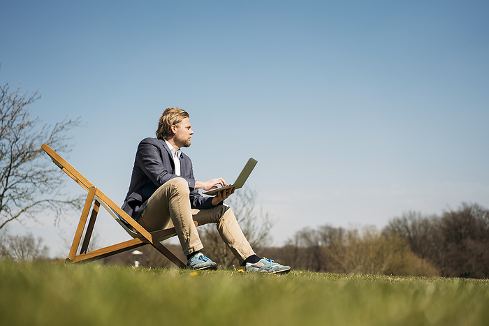 Surface level view of businessman looking away while sitting with laptop on chair at park during sunny summer day
