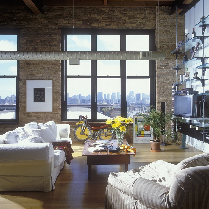 LIVING ROOM - Loft living with Chicago skyline, tall windows, wood floors, exposed brick walls, white sofa and chair, country style coffee table, palm palnt, toy motorbike, vase of spider mums, suspended shelving unit holds tv and media, heating duct